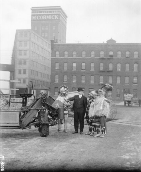 Man wearing a suit and a bowler hat is demonstrating the machine to a group of men in traditional Native American costume. There are factory buildings in the background.