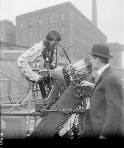 Man in bowler hat demonstrating machine to a man dressed in traditional Native American costume. They are in the factory yard.