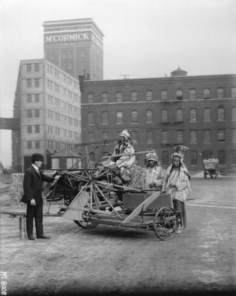 Man wearing a suit and a bowler hat is demonstrating the machine to a group of men in traditional Native American costume. There are factory buildings in the background.