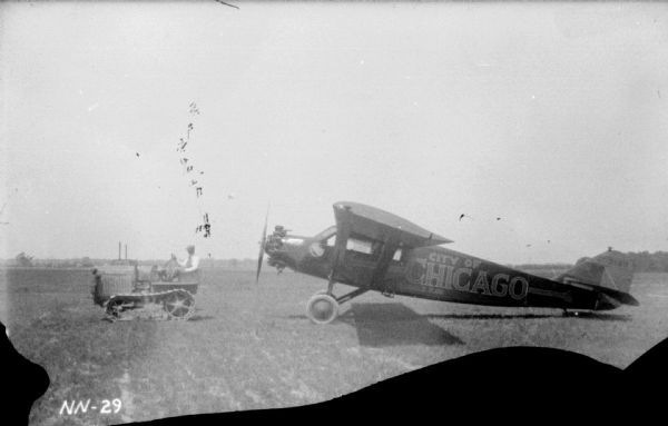 Left side view of a man driving a tractor with a continuous tracks wheels in front of an airplane outdoors in a field. The sign on the side of the airplane reads: "City of Chicago."