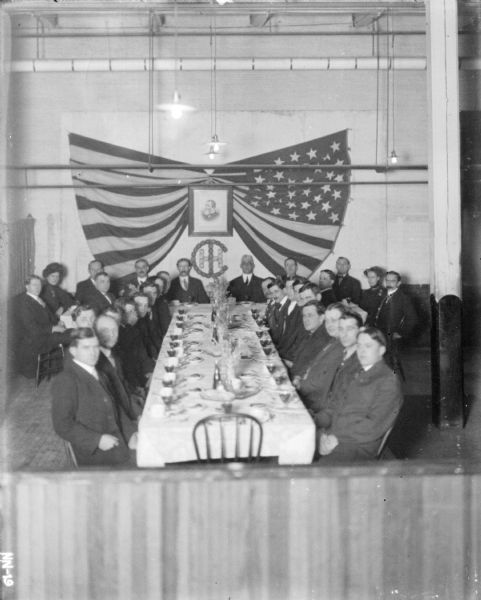 Group of people sitting around and near a table in a room decorated with banners. There is a portrait of Cyrus Hall McCormick, and the IHC logo below it, on the far wall.