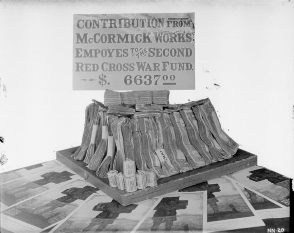Pile of money on top of posters, with a sign that reads: "Contribution from McCormick Works Employees, to the Second Red Cross War Fund $6637.00." The poster artwork is by artist McClelland Barclay and depicts the image of a fallen soldier being carried atop another soldier’s shoulders.