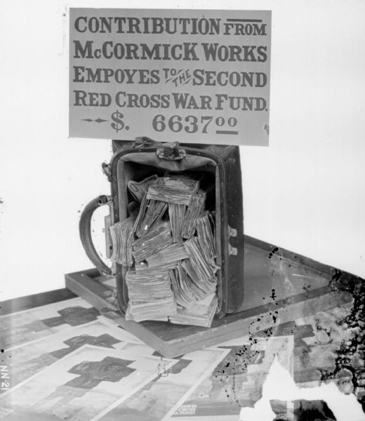 Pile of money in an open valise on top of posters, with a sign that reads: "Contribution from McCormick Works Employees, to the Second Red Cross War Fund $6637.00." The poster artwork is by artist McClelland Barclay and depicts the image of a fallen soldier being carried atop another soldier’s shoulders.