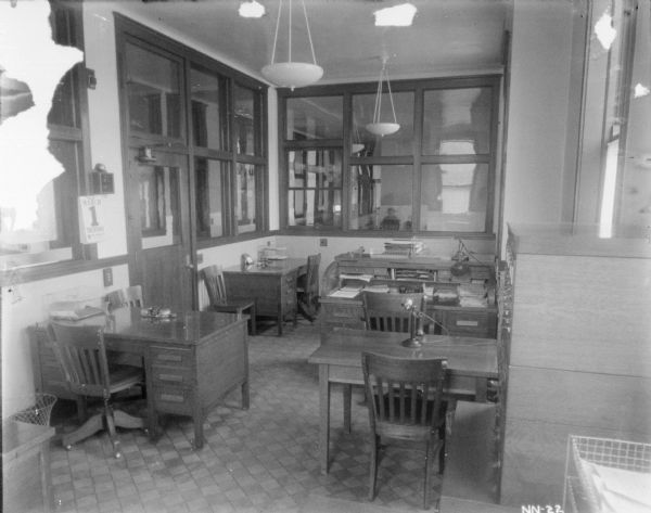 Office interior, showing a number of desks, including a roll top desk. There is telephone on one of the desks, and interior and exterior windows are around the room.