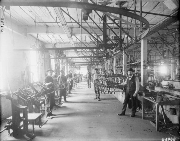 Men are standing at work stations on either side of an aisle in a large factory. There are large tracks or pulleys hanging from the ceiling. To the far right is a work table with tools. The men appear to be assembling pieces of farm machinery, and are all dressed in work clothes and hats, except the first man on the right who is wearing dress pants, a vest, a bow tie, and a hat. These workers are likely employees of the International Harvester Osbourne branch in Auburn, New York.