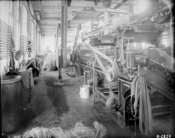 Factory floor, with a large piece of manufacturing machinery on the right, with other pieces of machinery directly behind it. The machine has many belts, pulleys, and gears. Hanging from the end of the machine in the foreground is an apron. The rest of the room is filled with bits of debris and large buckets, some filled with rags and brooms. The factory is likely the International Harvester Osbourne branch in Auburn, New York.