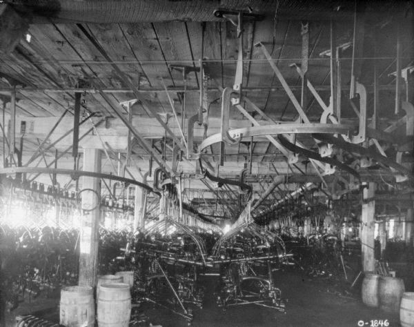 A large room in the factory is filled with pieces of farming implements in various stages of manufacturing. The parts are hanging from large hooks installed on tracks suspended from the ceiling. Scattered around the room are clumps of wooden barrels. This factory is likely the International Harvester Osbourne branch in Auburn, New York.