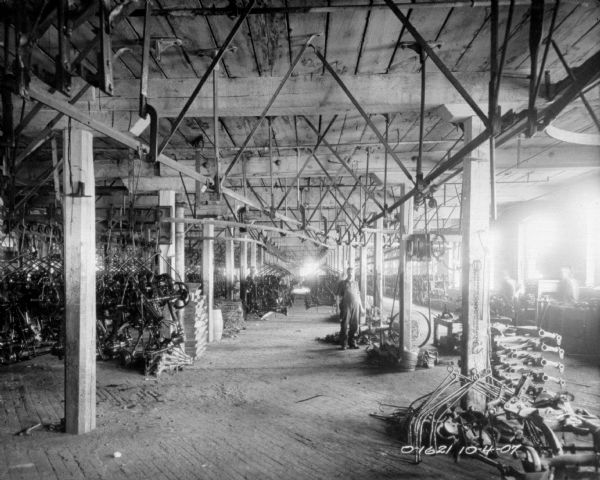 A man is standing in the middle aisle of a factory floor. To either side of him are stacked implement parts. To the right two men are at work stations. All three men are wearing work clothes, and they are probably employees of the International Harvester Osbourne branch in Auburn, New York.