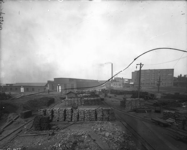 Elevated view of lumberyard. Men are standing in the yard, and buildings and smokestacks are in the background.
