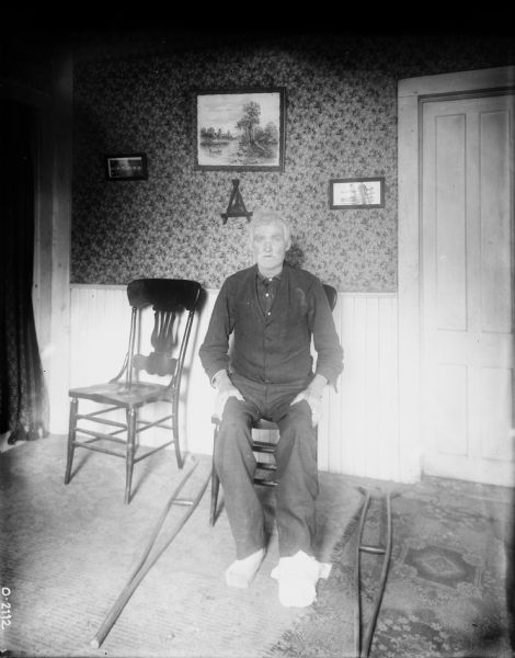 A man is sitting indoors in a room on a chair, with his left foot bandaged. Crutches are on the floor beside him. 