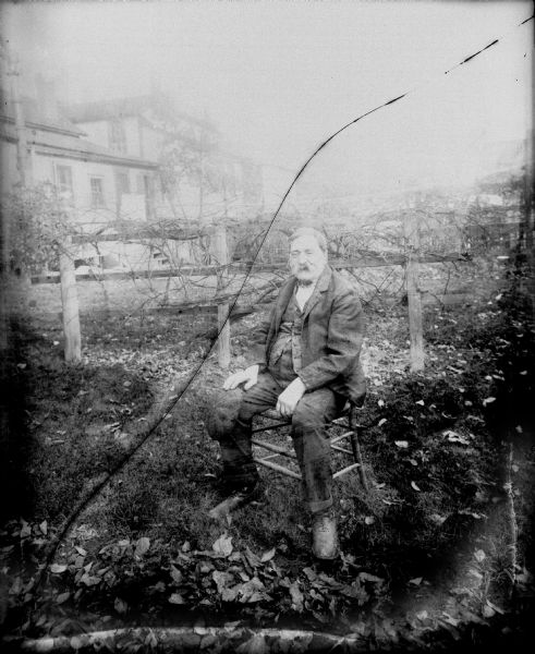 Man sitting on a chair in a backyard, holding a hat in his lap. Behind him are vines on a fence, and a house.