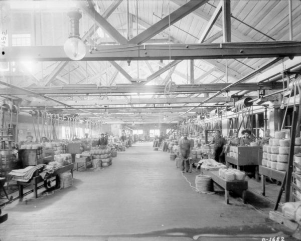 A view down the center aisle of what is most likely the International Harvester Osbourne factory in Auburn, New York. On either side of the aisle are men and women producing twine, probably for twine balers. The factory floor is open and bright with many windows, there are metal rafters hanging from the ceiling. The men and women are wearing work clothes, and one woman is wearing a large puffy hat.