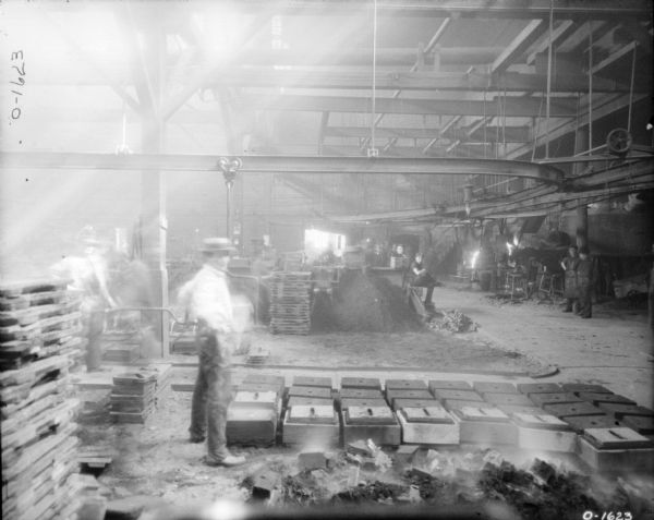 Piles of building materials and pallets are stacked inside the International Harvester Osbourne branch in Auburn, New York. There is a track hanging from the rafters of the ceiling, possibly for transporting materials. To the left are two men dressed in dark pants, button up shirts, and hats. To the left are groups of men dressed in work clothes and aprons.
