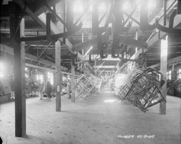 A view of the center aisle of a factory, (painting room?), with large windows, wooden rafters, and a wooden floor. Hanging from the ceiling from large hooks are what appear to be frames to trucks. To the left are three men. The factory is likely located at the International Harvester Osbourne branch in Auburn, New York.