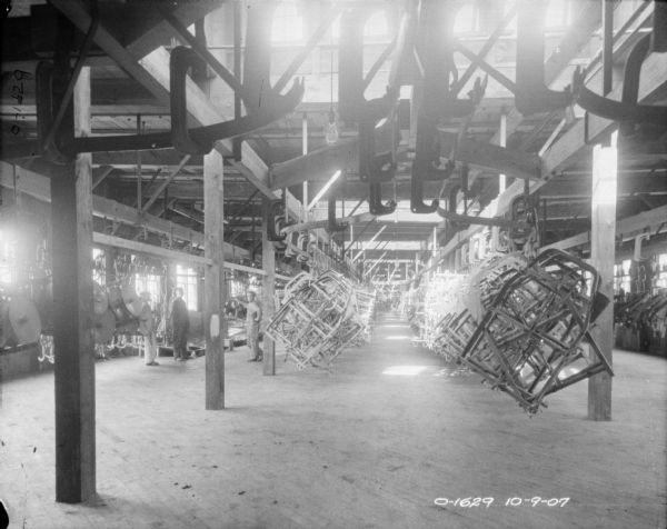 A view of the center aisle of a factory, (painting room?), with large windows, wooden rafters, and a wooden floor. Hanging from the ceiling from large hooks are what appear to be frames to trucks. To the left are three men. The factory is likely located at the International Harvester Osbourne branch in Auburn, New York.