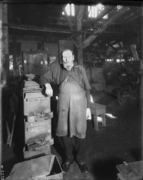 A man wearing an apron is standing and leaning on forms near a workbench.