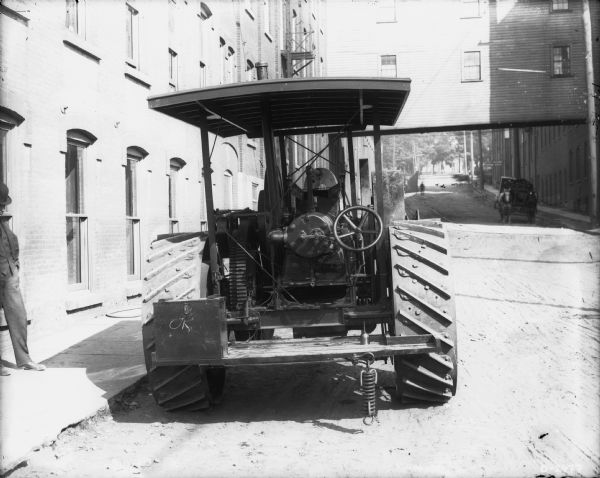Rear view of a Steam Tractor parked outdoors near factory buildings. A man is standing on the far left. A horse-drawn wagon is in the background on the right under a sky bridge between two buildings.