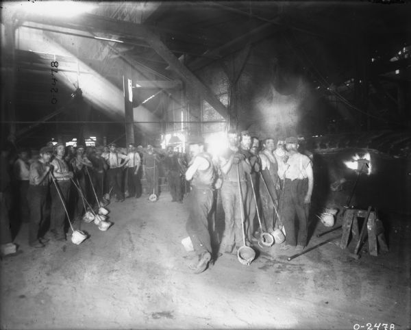 A group of male employees are posing, leaning on tools, in foundry area. There is a skylight pouring light down into the room.
