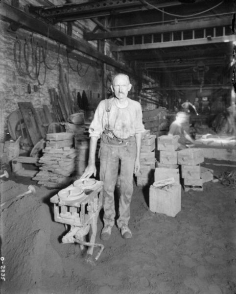 A man is standing on a factory floor. He is wearing overalls, a belt, and a loose shirt. Behind him are other men and assorted building materials. The factory floor looks to be made of dirt. The men are likely employees of the International Harvester Osbourne branch in Auburn, New York.