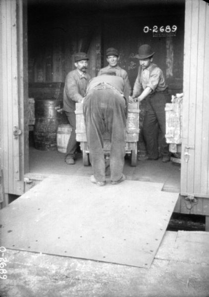 Four men are transporting a cart of goods from a railroad car to a loading dock. The men are most likely employees of the International Harvester Osbourne branch, located in Auburn, New York. In the background are barrels and other carts. The four men are wearing dark coats, collared shirts, and hats. Two of the men have moustaches.
