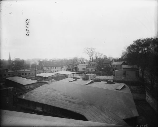 Elevated view over roofs of buildings of a factory. Wheels are stacked up in the yard. Includes a view of the neighborhood beyond.