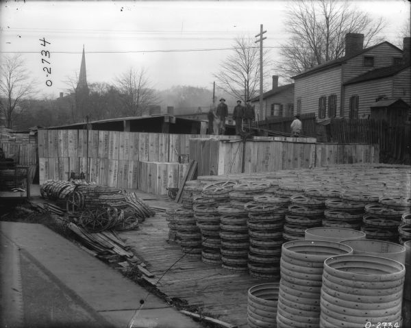 Wheels are stacked in factory yard, and men are standing on a platform behind. There is a view of a neighborhood in the background.