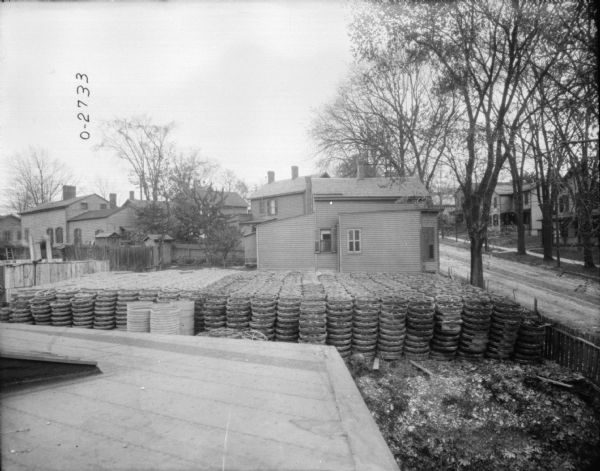 Elevated view of several rows of stacked wheels that are in the yard of what may be the International Harvester Osbourne branch in Auburn, New York. Next to the stack is a wooden platform. In the background on the right is a street and a neighborhood of wooden homes.