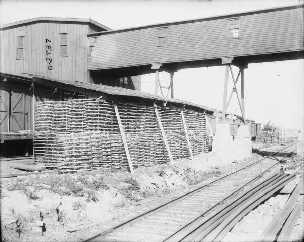 Wheels stored outdoors under a slanted roof near a factory building. Railroad tracks are in the foreground, and a building with a loading dock and a skybridge is on the right. 