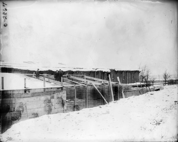 Three men are standing on top of stacks of wheels, constructing a roof over a storage area. Factory buildings with a loading dock are behind them, and snow is on the ground.