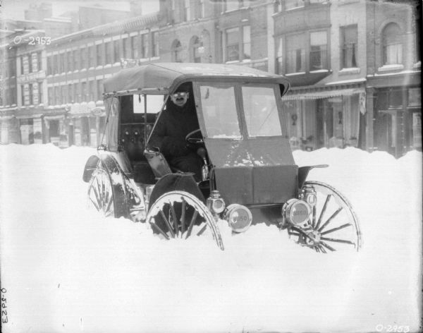 View towards a man driving a car in deep snow on a street. Commercial buildings are in the background. The driver's seat is on the right side, and the sides of the car are open. Numbers on the headlights of the car read: "81150."