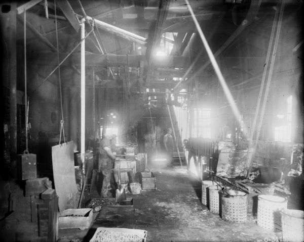 Men are standing in a manufacturing area with an exposed ceiling. Machinery in the room is belt-driven from the ceiling. The man on the left is holding a shovel. Barrels and perforated metal pails are on the floor throughout the room.