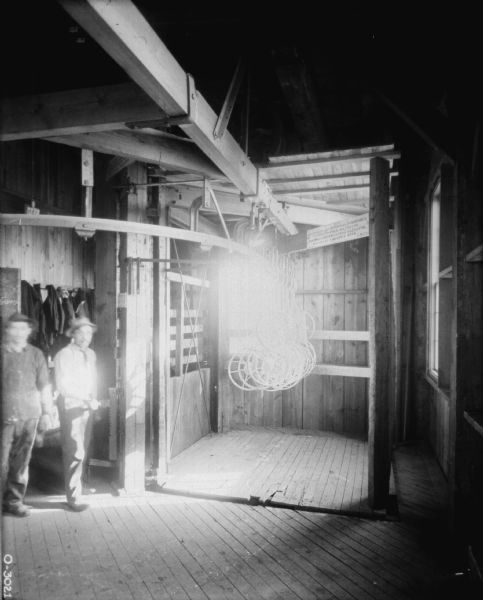 Two men dressed in work clothes are standing next to what appears to be an elevator. In the elevator there are several implement parts hanging from the ceiling, and there is a sign above that is difficult to read, but mentions something about "risk" and was posted by "I.H.C." The room is lit by two windows with bright sunlight shining in. This photograph may have been taken at International Harvester Osbourne Work in Auburn, New York.