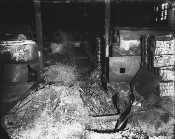 Interior view of a cluttered manufacturing area. There is a barrel on the left, and metal parts are in a pit in the center. A man blurred by movement is in the background.
