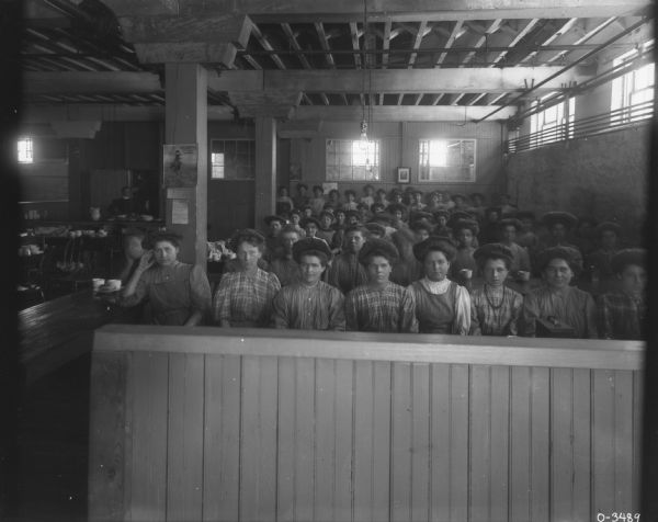 A large group of women employees are posing and sitting in rows in the dining room. They are all wearing hats.