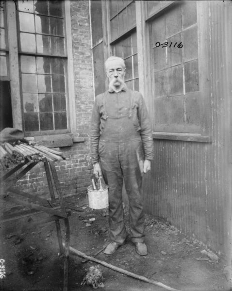 A man dressed in overalls and a button-up shirt standing in the corner of a factory yard in front of a brick wall and a corrugated wall with windows. The man appears to be holding a bucket of paint, with a paint brush in one hand and something flat in the other. Next to the man is makeshift work table with lumber. This man may be an employee of the International Harvester Osbourne branch in Auburn, New York.
