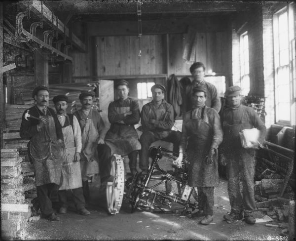 Group portrait of men standing on a factory floor together around a piece of agricultural machinery. Some of the men are holding hammers, and wearing aprons.