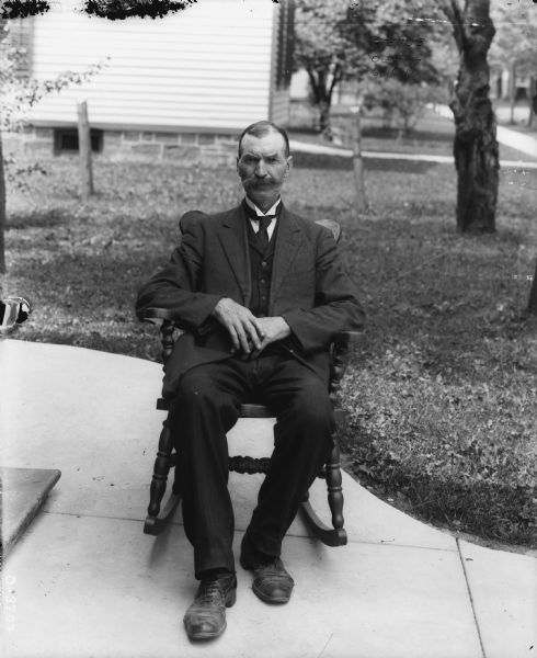 Portrait of a man sitting in a chair on a sidewalk near a building. In the background are trees in a lawn, and another building.