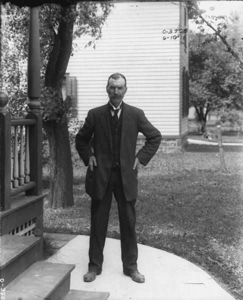Portrait of a man standing on a sidewalk near the porch of a building. In the background are trees in a lawn, and another building.
