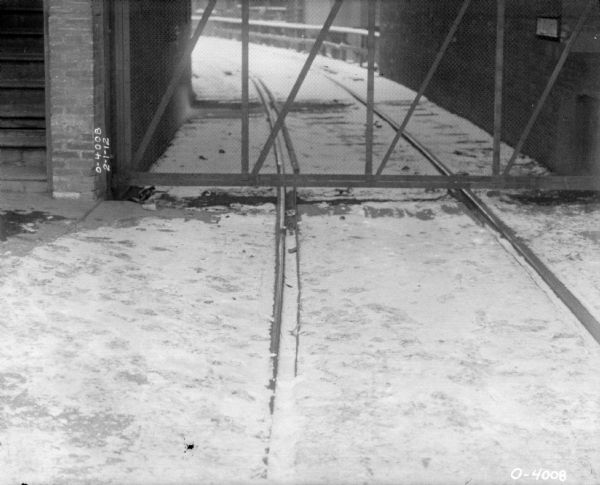 Railroad tracks covered in snow are curving under a gate leading into a factory yard. The factory is likely the International Harvester Osbourne branch in Auburn, New York.