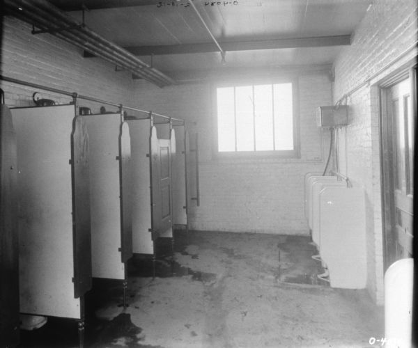 This photograph may be of the men's bathroom at the International Harvester Osbourne branch in Auburn, New York. To the right are several urinals, while on the left are several bathroom stalls. The floor is wet and dirty. There are pipes on the ceiling, a window to the back of the room, and a door to the right.