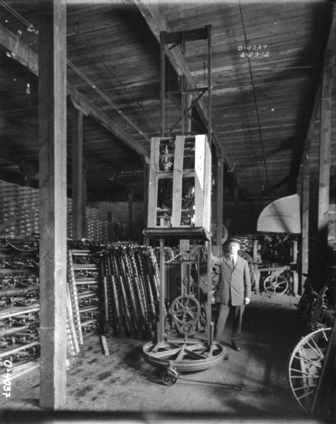 A man dressed in a suit and hat is standing next to a crane lifting machine crated for delivery. The wood building has tall ceilings and is filled with stacks of implement parts. Behind the man is an awning that reads: "Osbourne." This man is likely an employee of the International Harvester Osbourne Works in Auburn, New York.
