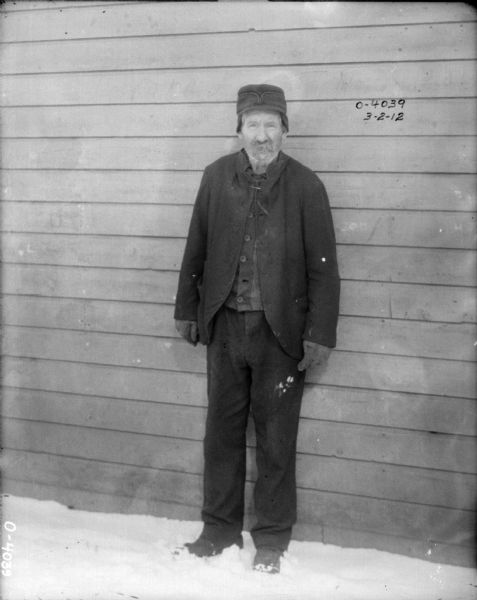 Portrait of a man standing in the snow against a wood-sided building. He is warmly dressed in a coat, hat, shoes and gloves.
