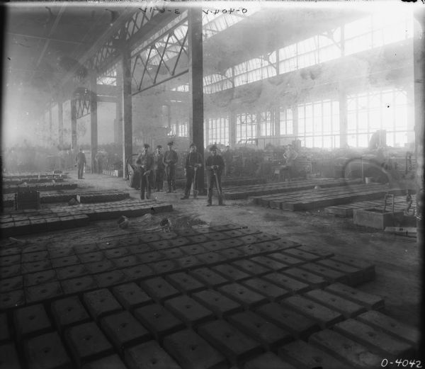 Men are posing in a foundry area with a high ceiling and skylights. Metal forms are laid out in rows on the floor, and many of the men are holding pitchforks. A large row of windows is along the back wall.