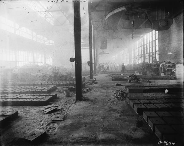 Men are posing in a foundry area with a high ceiling and skylights. Metal forms are laid out in rows on the floor, and many of the men are holding pitchforks. A man is in the cab of a crane or conveying machinery which is suspended from a track that runs in a curve above the metal forms. A large row of windows is along the back wall.