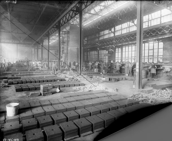 Men are posing in a foundry area with a high ceiling and skylights, and a tall, brick furnace is in the center. Metal forms are laid out in rows on the floor in the foreground, and many of the men are holding shovels or pitchforks. A man is in the cab of a crane or conveying machinery which is suspended from a track near the furnace. A large row of windows is along the back wall.