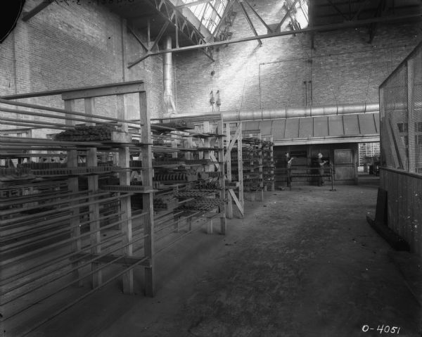 View across factory floor towards men standing in the background near metal shelves. Rows of metal shelves are along the left, and hold metal parts. There are skylights above in the exposed ceiling.