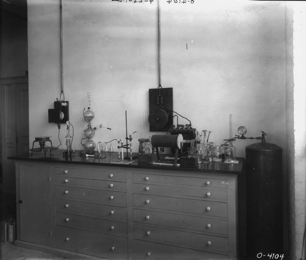 Interior view of a cabinet with doors and drawers against a wall, with various instruments and glass vessels arranged on the counter. There is a tank with a dial on the right set up to an instrument on the counter.