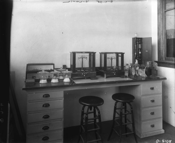 Interior view of a laboratory. Two stools are set up in front of a bench with drawers along the sides sitting against a wall. Various instruments and glass vessels are arranged on the counter, and small scales are inside two glass cabinets in the center of the counter.