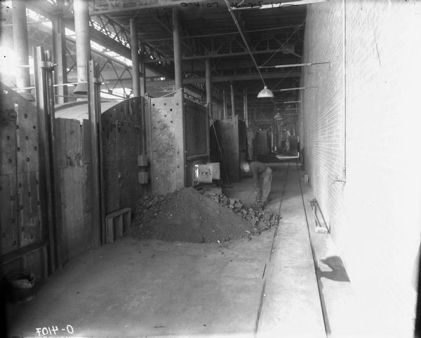 A man (blurred by movement) using a shovel is bending over a pile of coal near the open door of a furnace indoors on factory floor. More furnaces are in the background, and tracks are running along the floor.