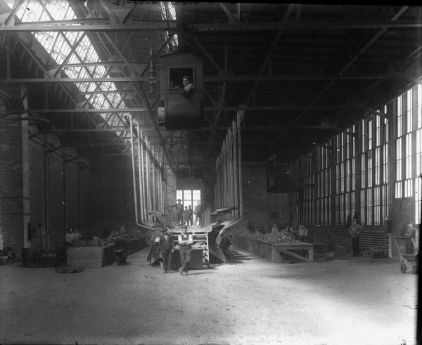 Men posing on factory floor in a metal finishing area. In the center is a man in the cab of a crane overhead. There are skylights in the ceiling on the left.